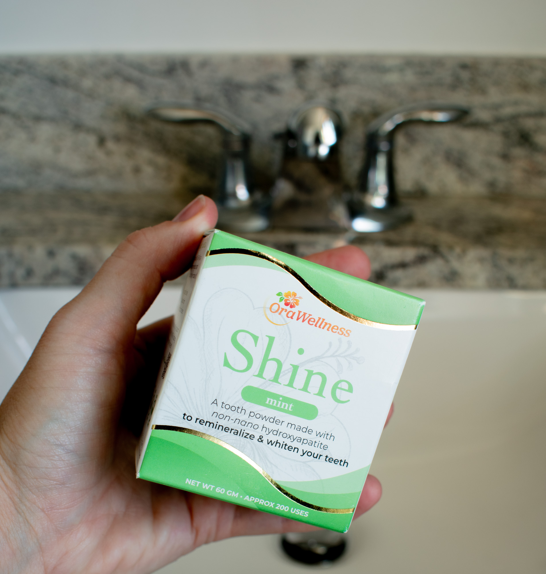 A person's hand holding a box of OraWellness Shine above a bathroom countertop